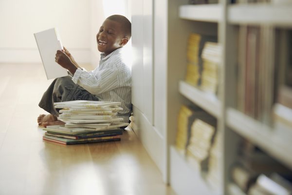 Image of a young boy laughing while reading a braille book and leaning against a library shelf.